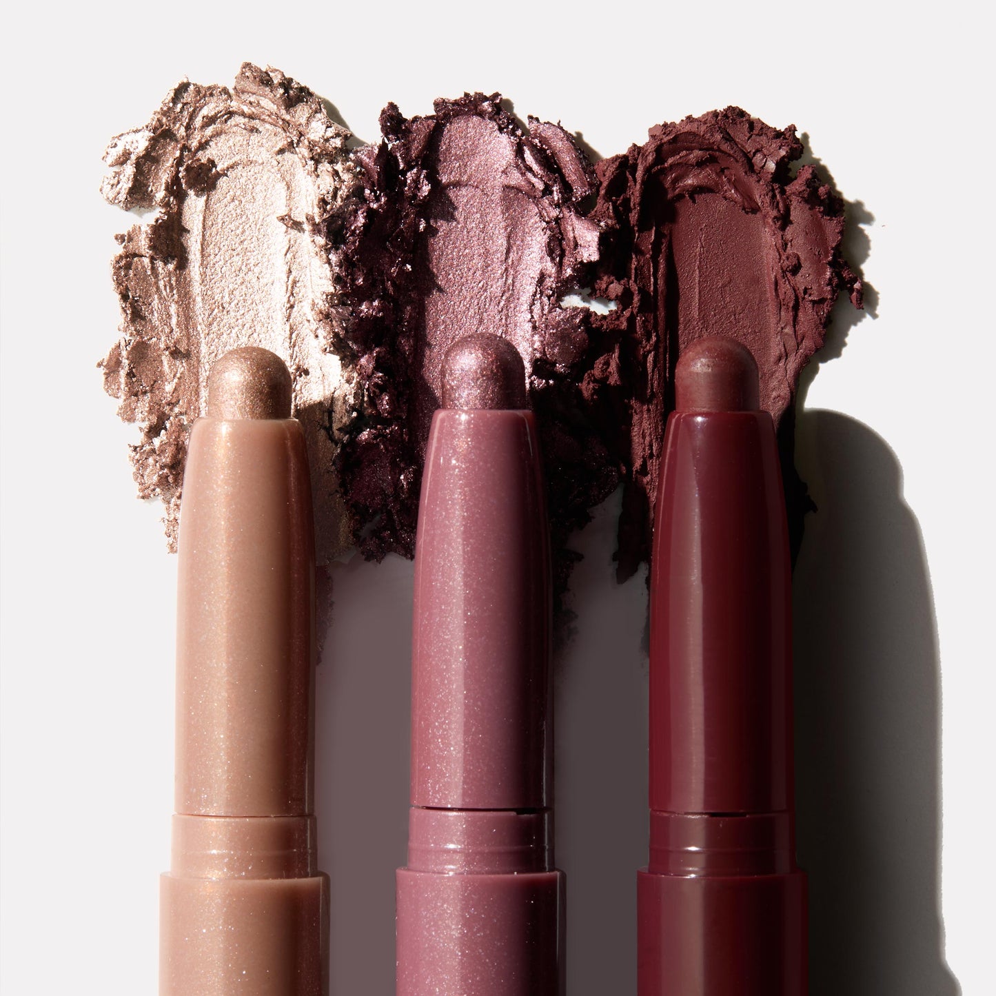 Three Rose Goals ColourPop Shadow Stix showcase creamy swatches - champagne shimmer, rosy mauve metallic, and deep burgundy - smeared behind the twist-up pencils on a white backdrop, highlighting the eyeshadow stick trio.