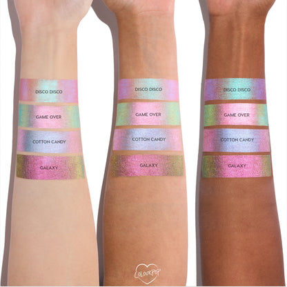 Colourpop Shadow Stix Game Over swatched on three skin tones, showing the shimmery pink-to-green duochrome strip labeled Game Over between Disco Disco and Cotton Candy shades on each forearm for comparison.